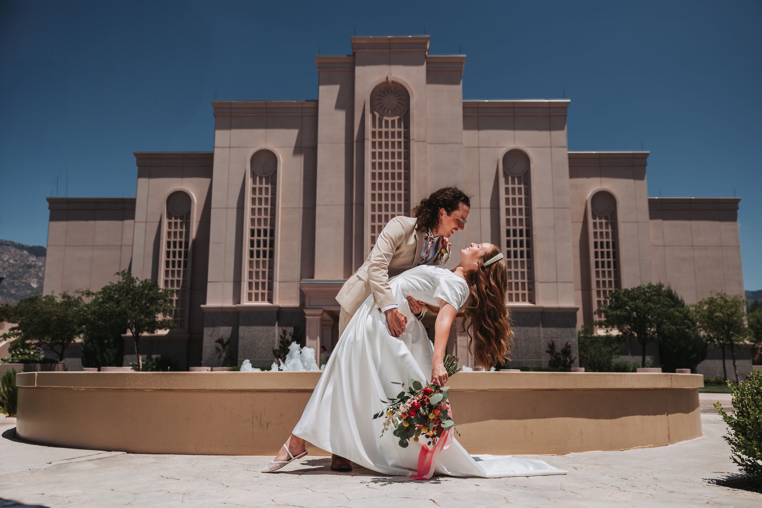 A romantic wedding couple poses in an elegant dip with a beige stone church facade and arched windows in the background.