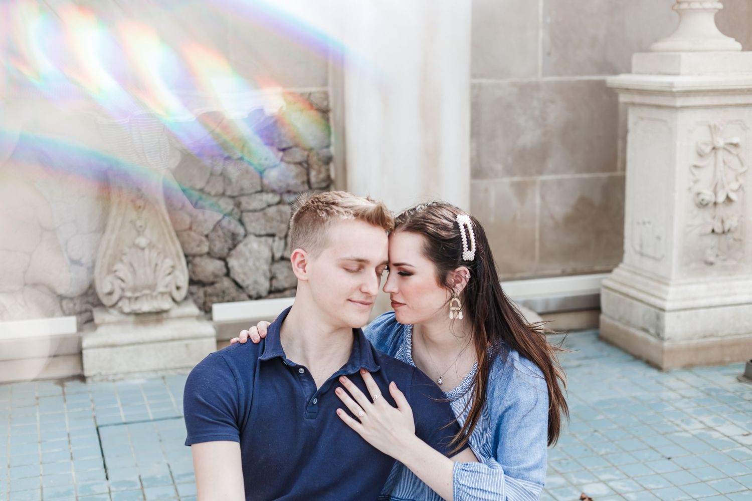 Two people share a tender romantic moment in an elegant outdoor setting with ornate stone architecture.