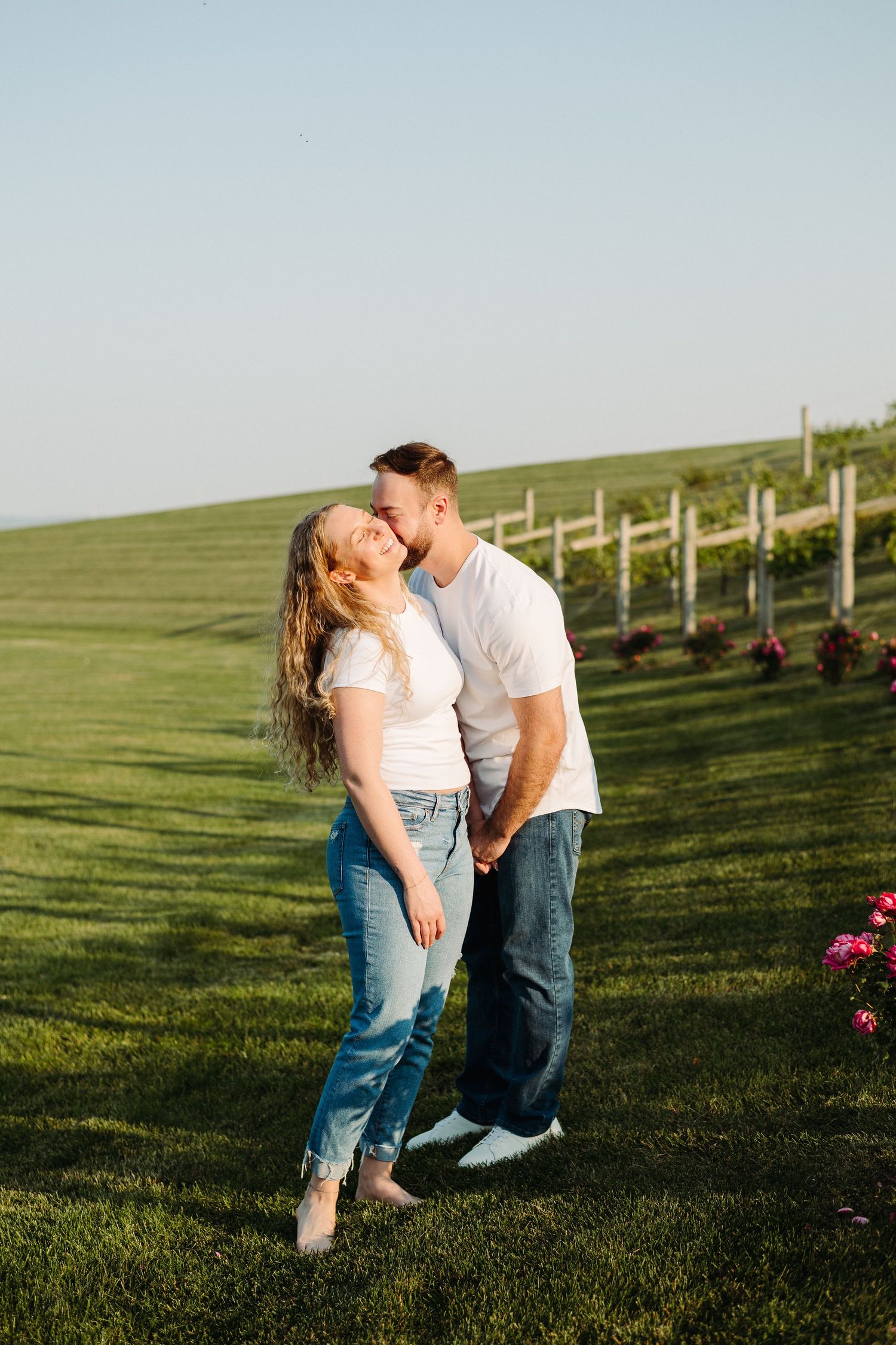 A couple shares a romantic moment in a green field with fencing during golden hour sunlight.