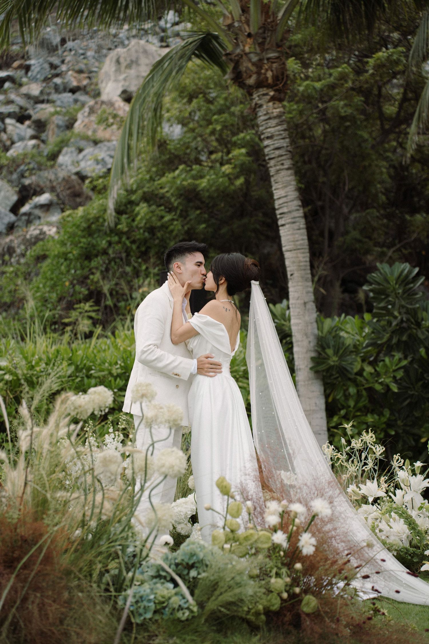 Couple in white attire embraces amid lush tropical garden setting with palm trees and natural greenery at golden hour.