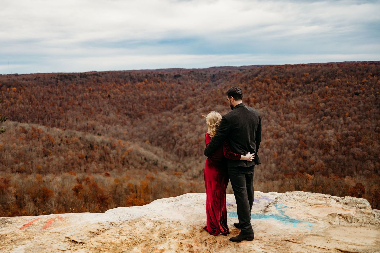 Couple in red dress and black suit embrace on rocky overlook with autumn forest.