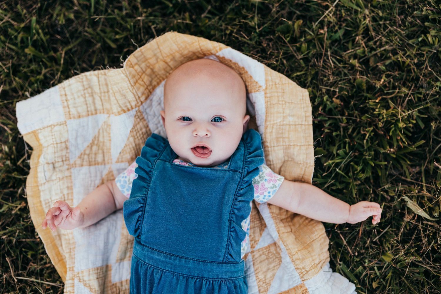 Baby in blue denim overalls plays on yellow quilt spread over green grass on sunny day.