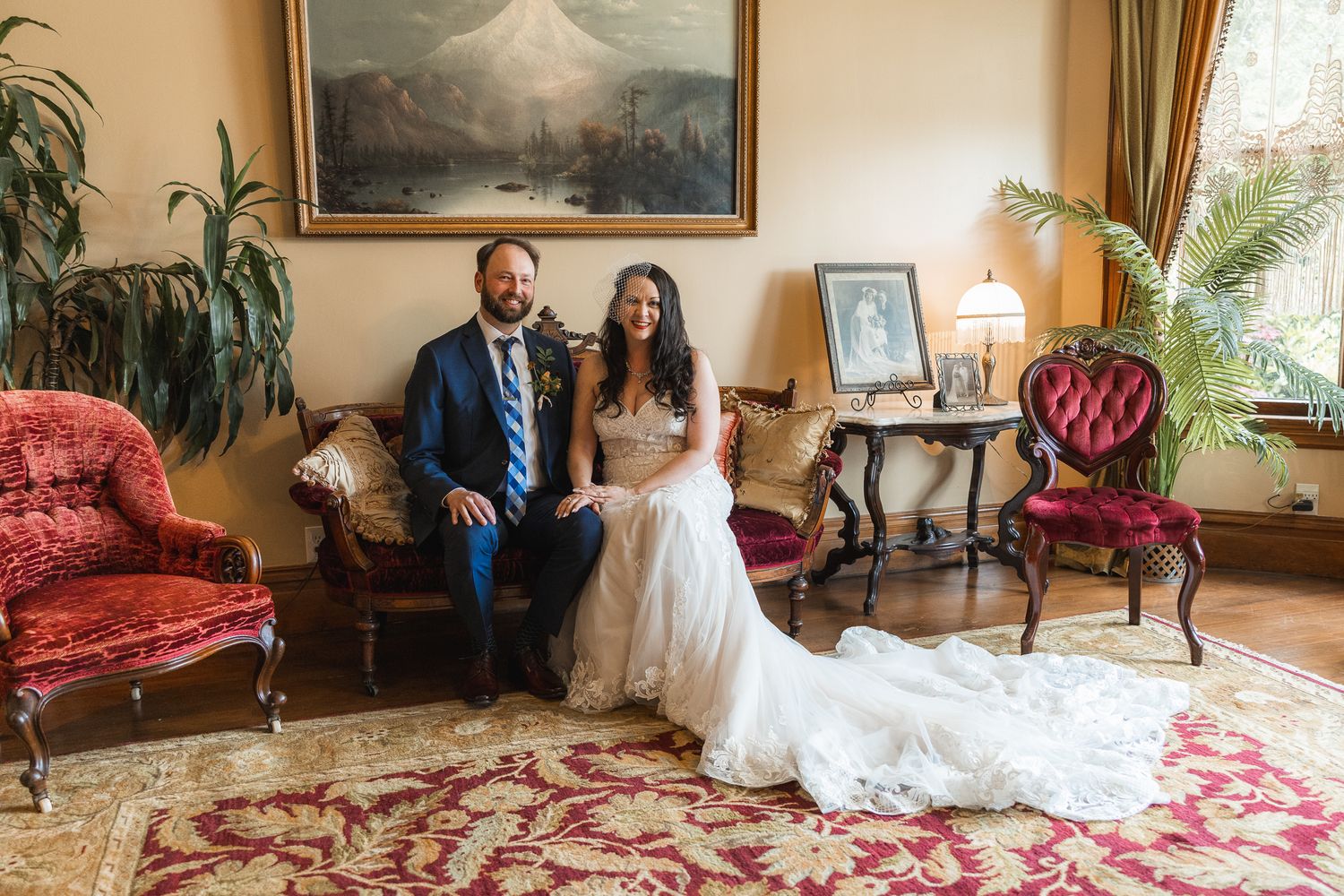 A wedding couple poses in an elegant Victorian-style room with ornate furniture and red floral decor.