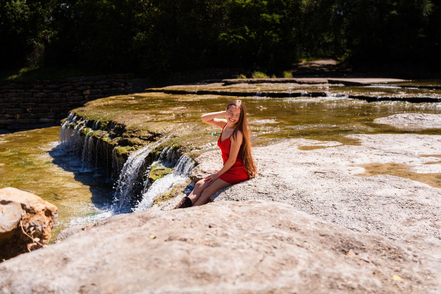 Two people sit together on rocks near a scenic tiered waterfall with lush greenery in the background.