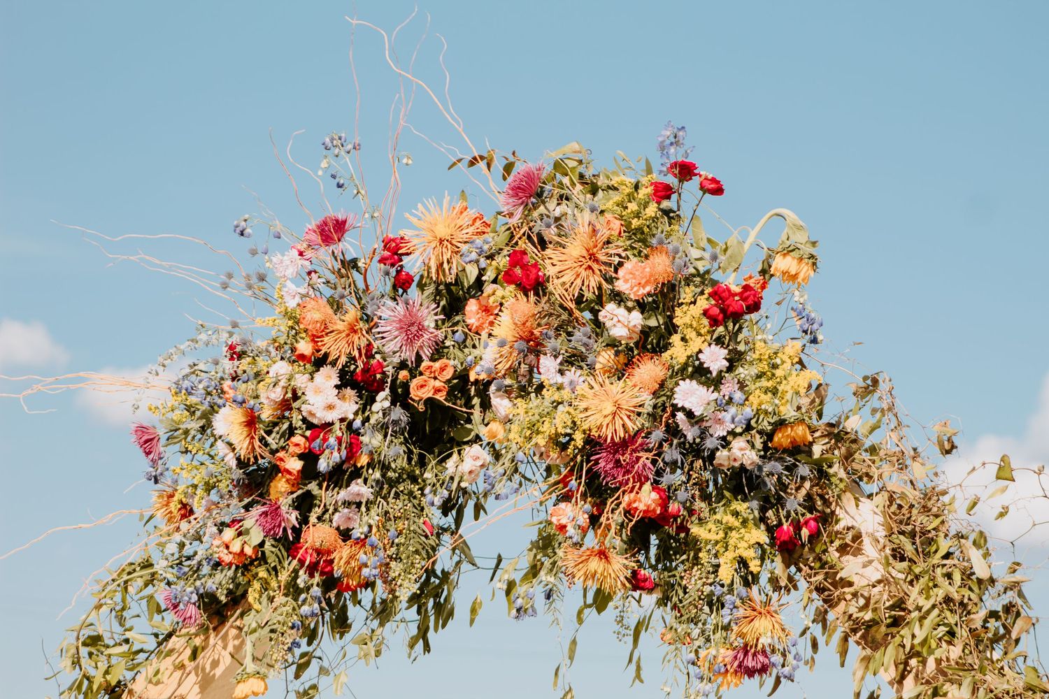 Colorful floral arch with dahlias, wildflowers, and greenery against a blue sky for a wedding ceremony.