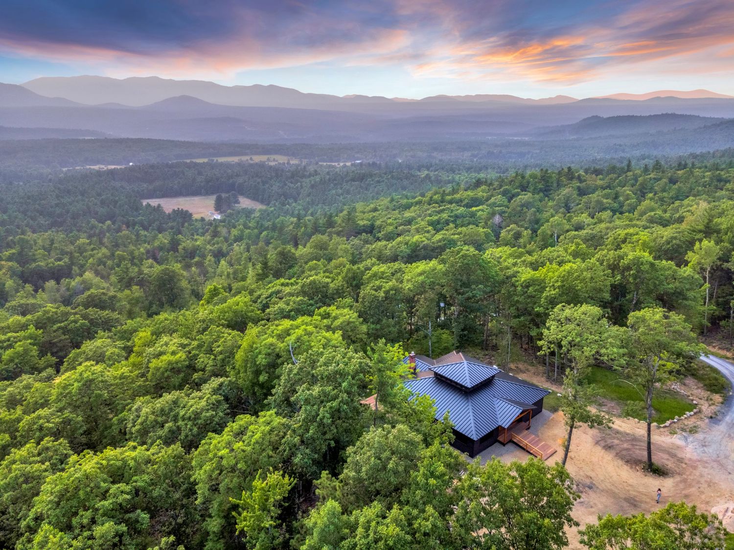 A serene panoramic mountain landscape with lush green forest canopy stretches to the horizon under a colorful sunset sky.