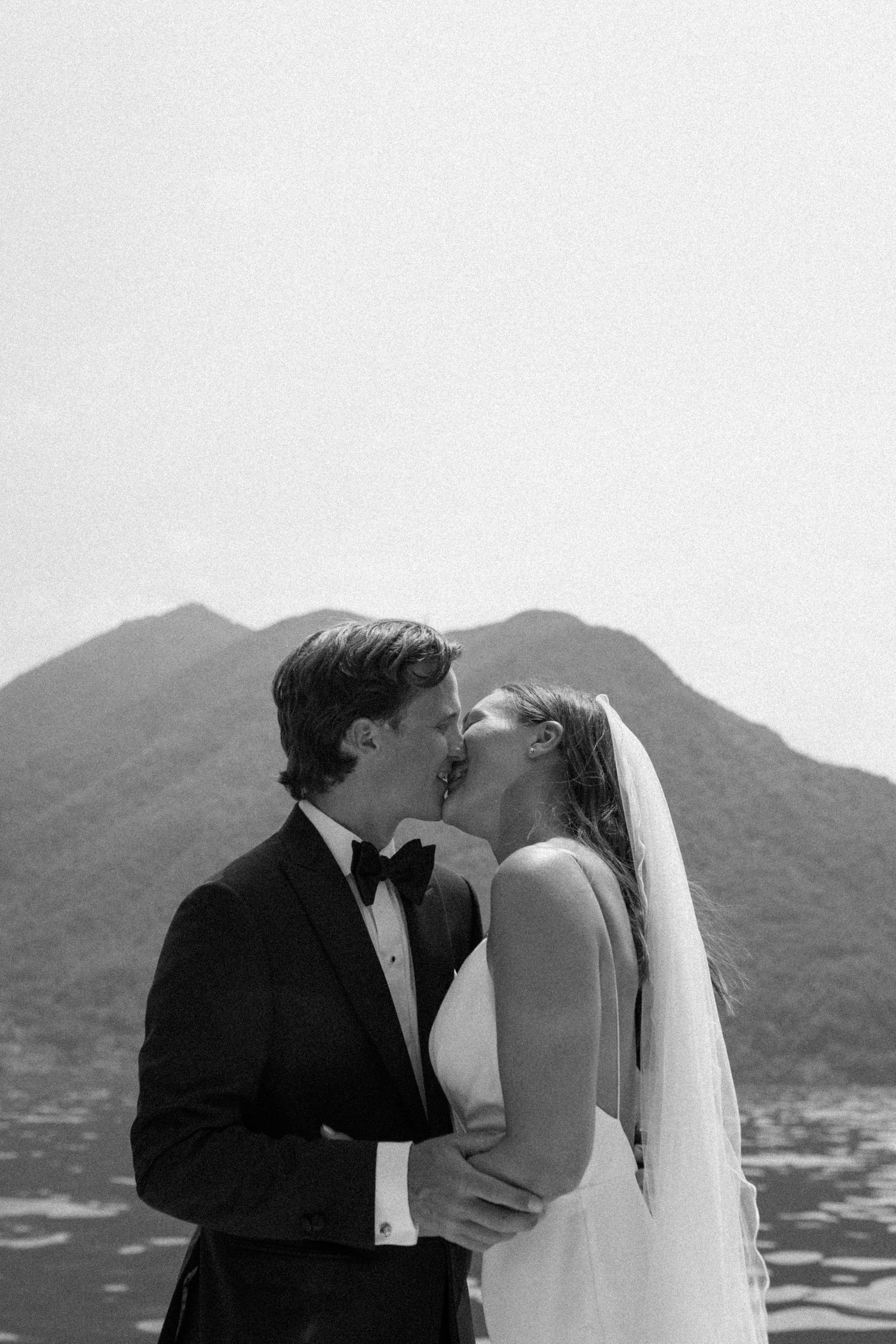 A bride and groom share a romantic kiss against a backdrop of misty mountains in black and white at their Lake Como wedding.