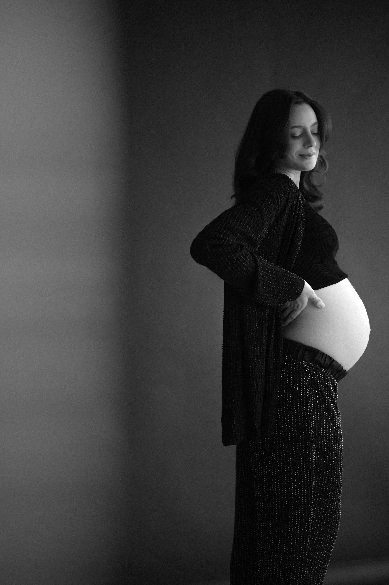 A dramatic black and white maternity photo sequence showing a woman in dark clothing against a shadowy background.