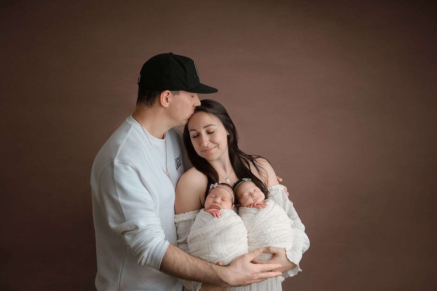 A tender family portrait showing parents gently cradling their newborn baby in a warm brown-toned studio setting.