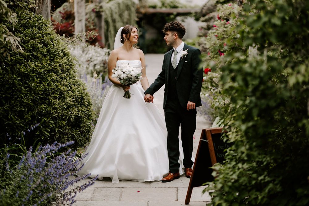 An elegant couple walking together amidst lush green foliage during their garden wedding ceremony.