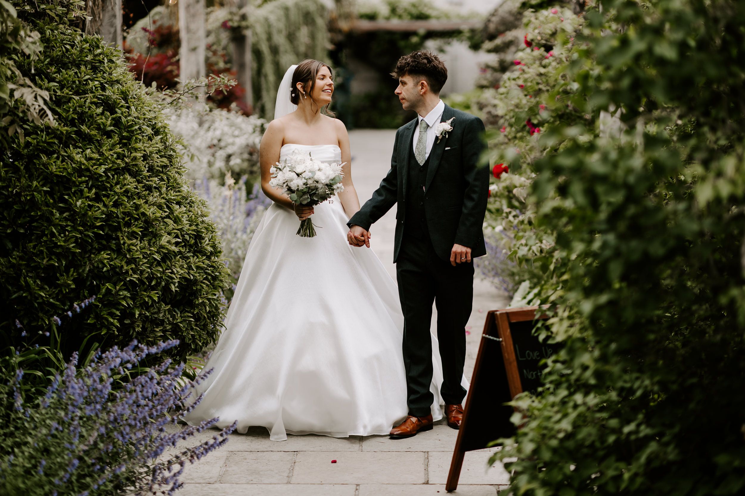 An elegant couple walking together amidst lush green foliage during their garden wedding ceremony.