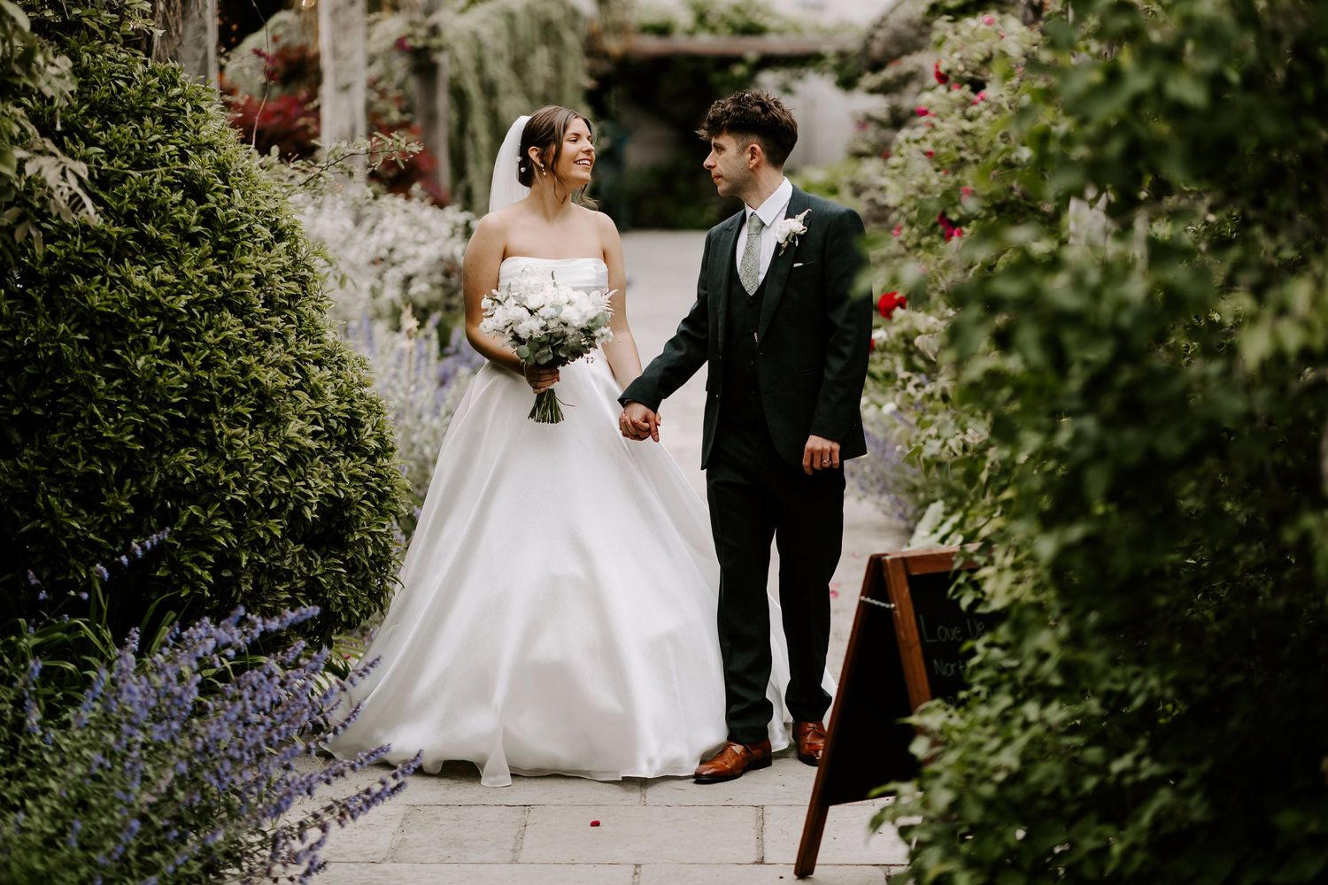 An elegant couple walking together amidst lush green foliage during their garden wedding ceremony.