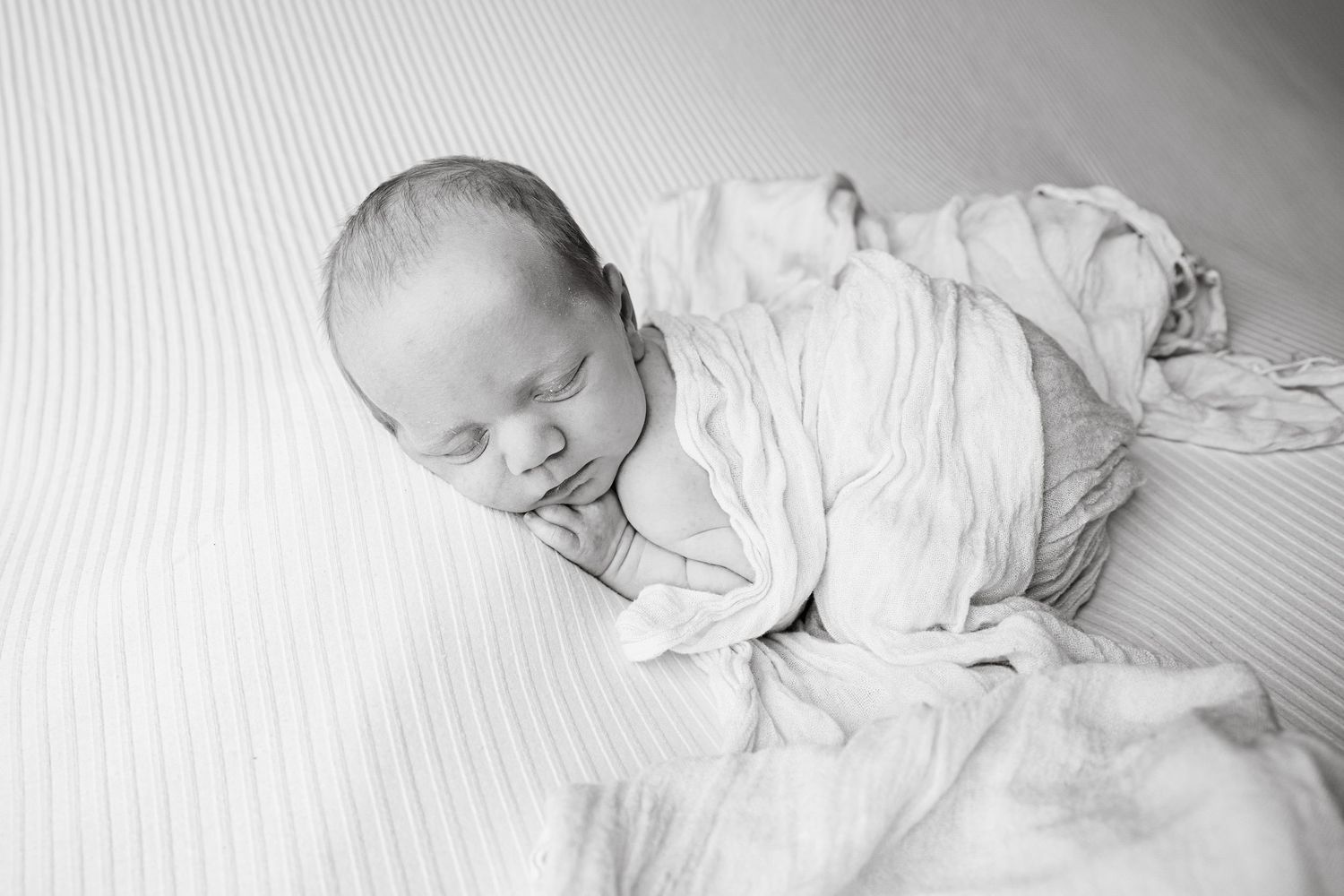 A sleeping newborn wearing white clothing lies on white bedding in black and white photography.