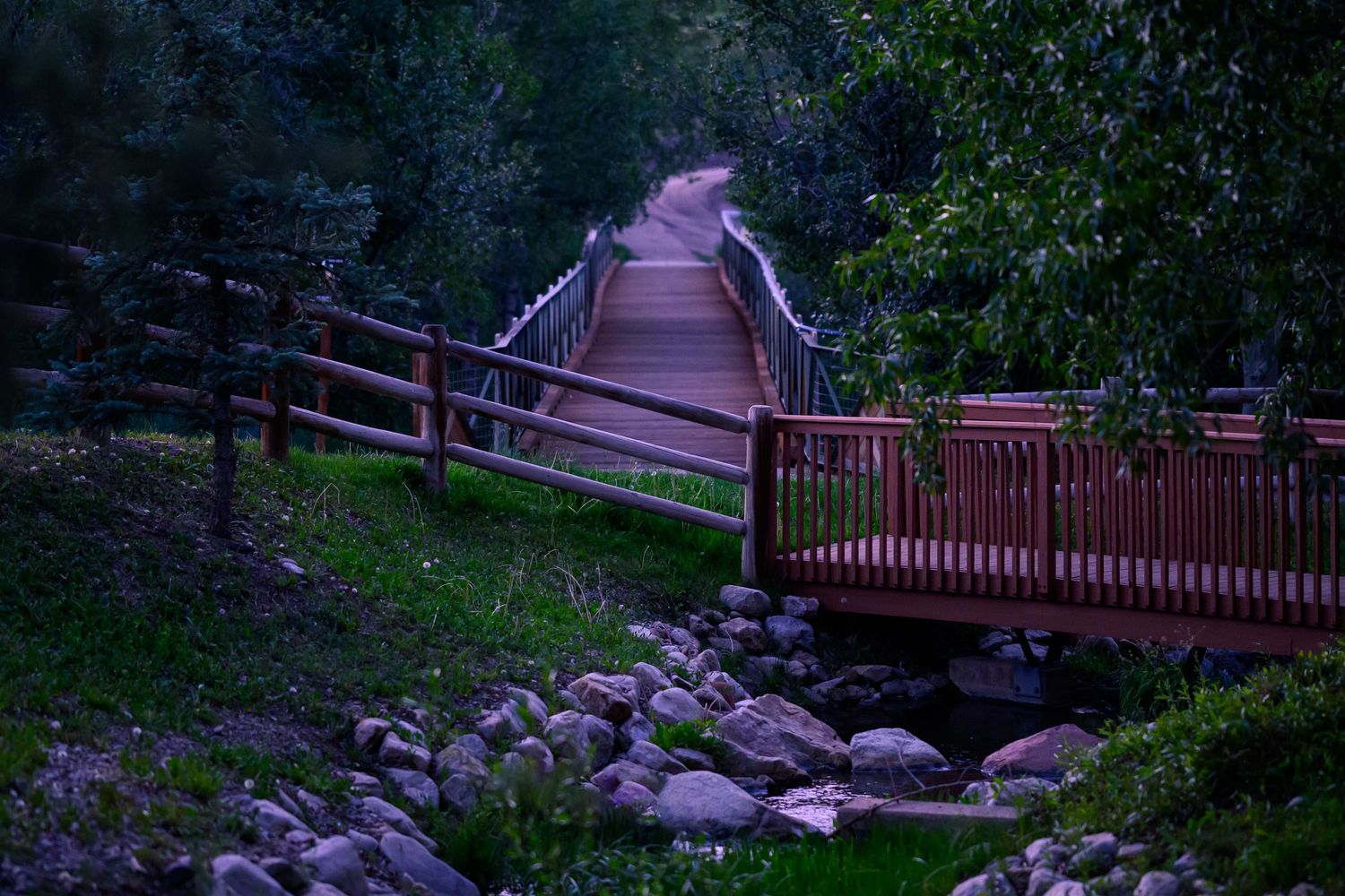 Wooden footbridge with red railings over a rocky stream surrounded by lush green trees at dusk.