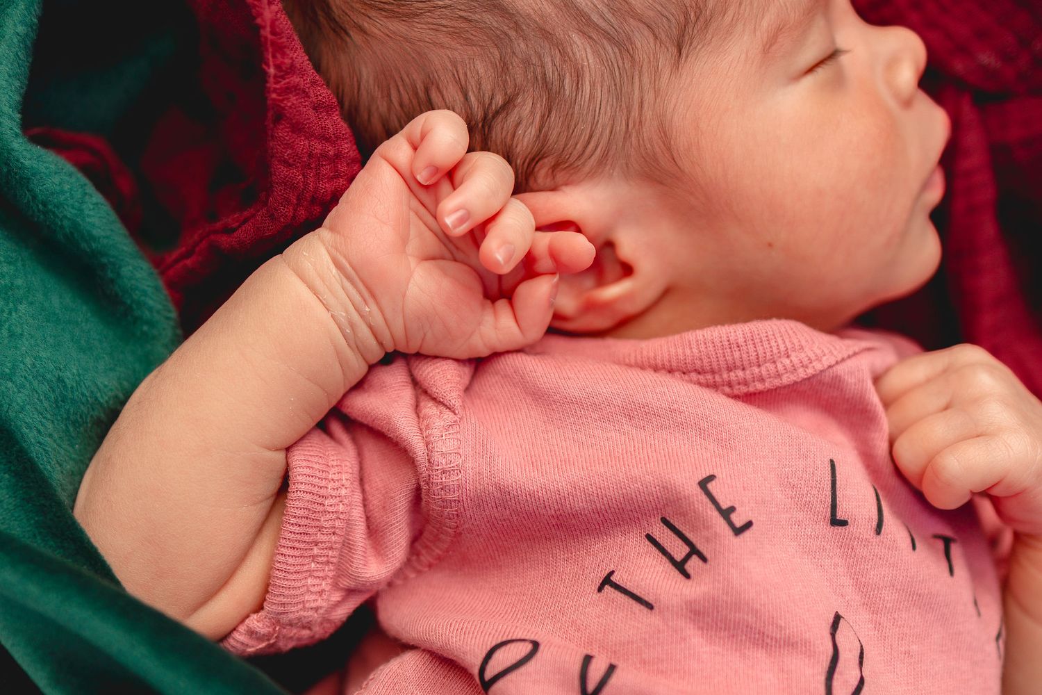 Sleeping newborn baby in pink onesie with tiny hand raised near ear, resting on colorful blankets.