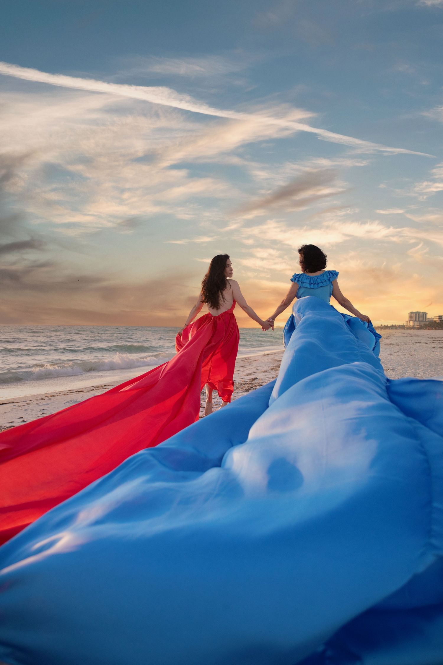 Two figures in flowing red and blue dresses dance together on a beach at sunset with dramatic clouds in the sky. Flying Dress Photoshoot