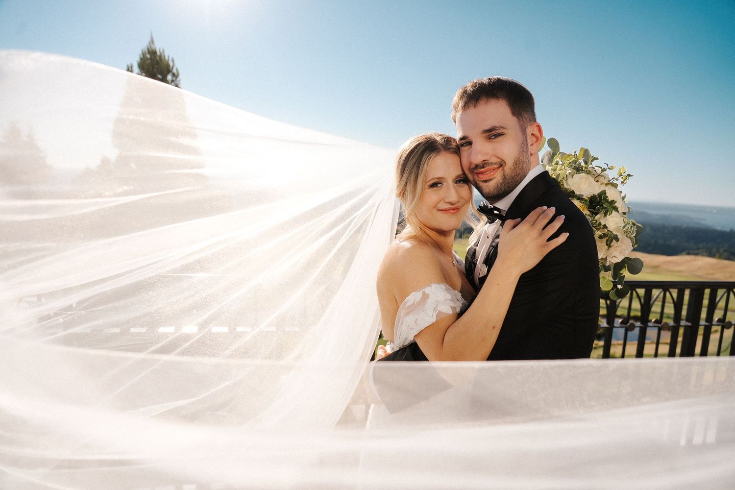 A couple embraces during their outdoor wedding ceremony as a veil flows gracefully in the breeze.