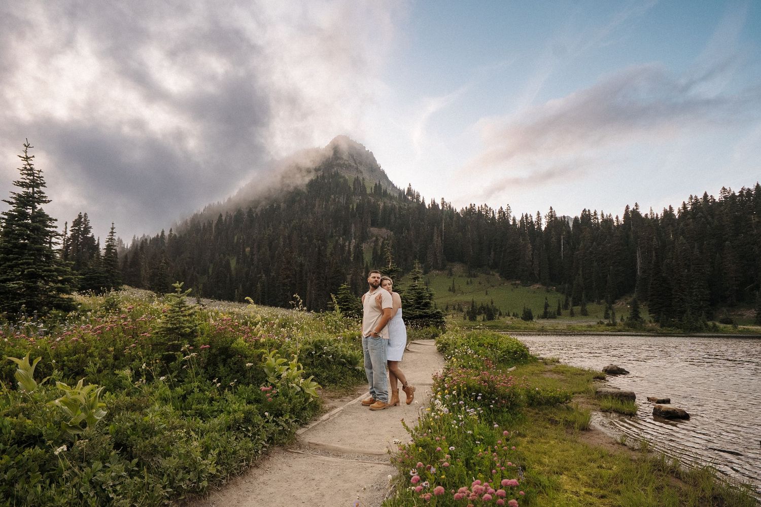 Two people walk along a mountain trail surrounded by wildflowers and evergreen trees beneath cloudy skies.