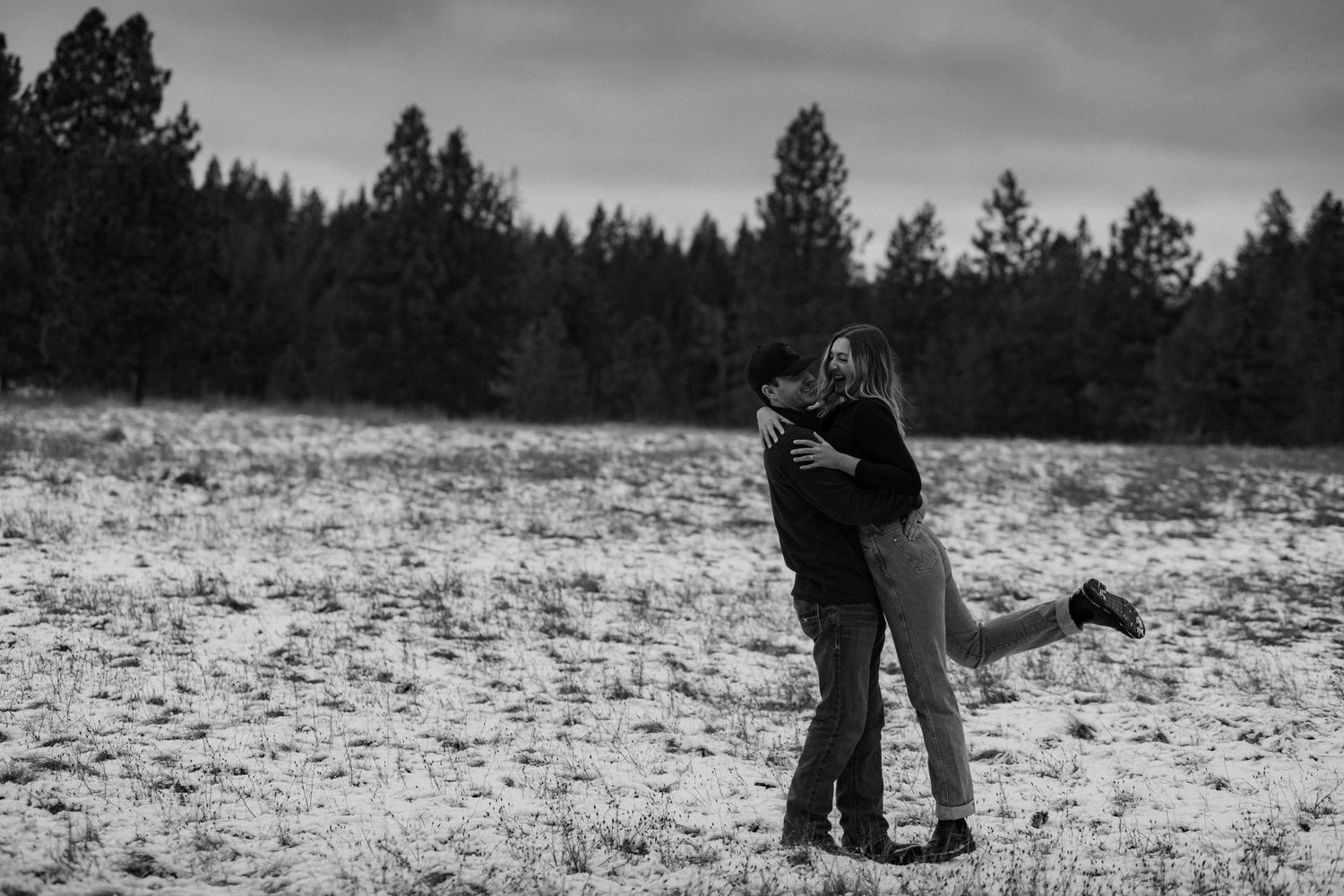 A couple embraces in a snowy winter landscape with evergreen trees in the background.