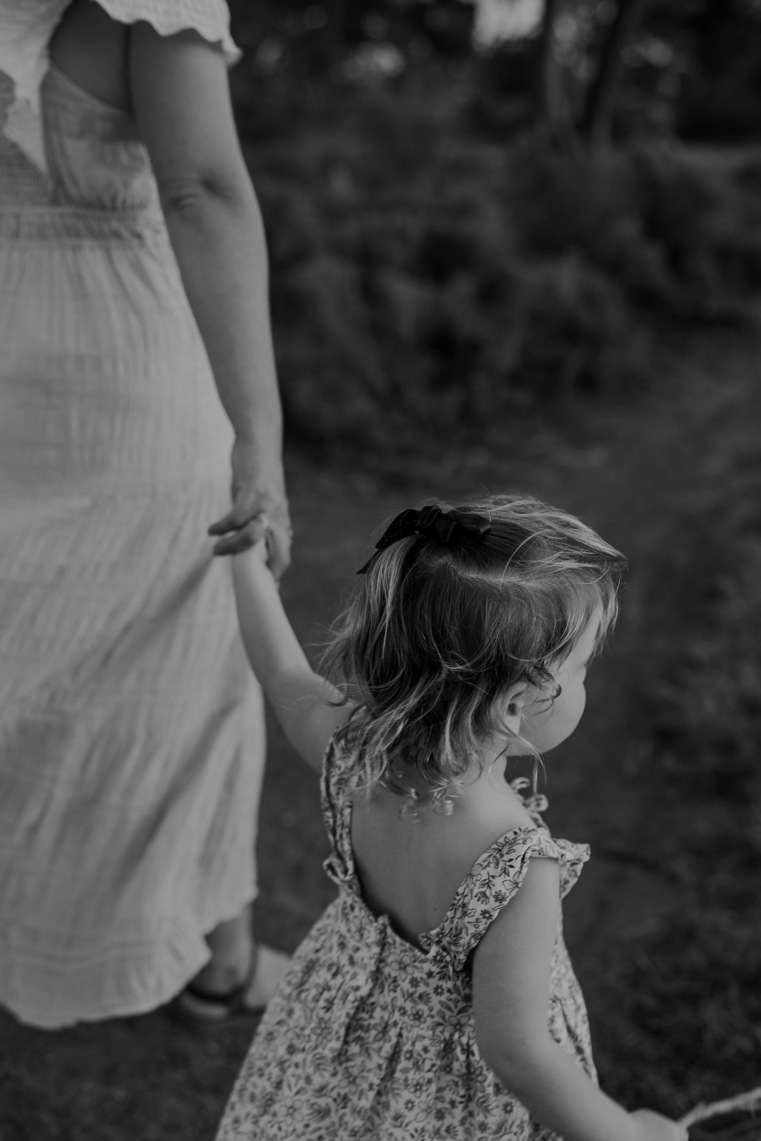 Black and white image showing a small child in a floral dress walking outdoors on a sunny day.