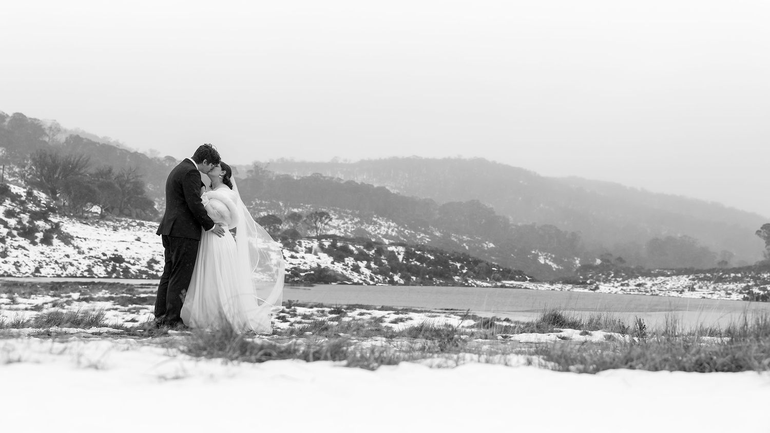 A couple in wedding attire embraces on a snowy hilltop overlooking a picturesque mountain landscape in black and white at Perisher.