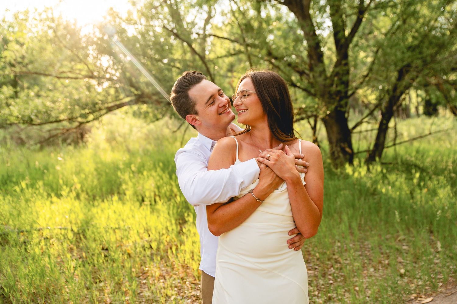 A couple shares a romantic embrace in a sunlit meadow with trees and yellow wildflowers in the background.