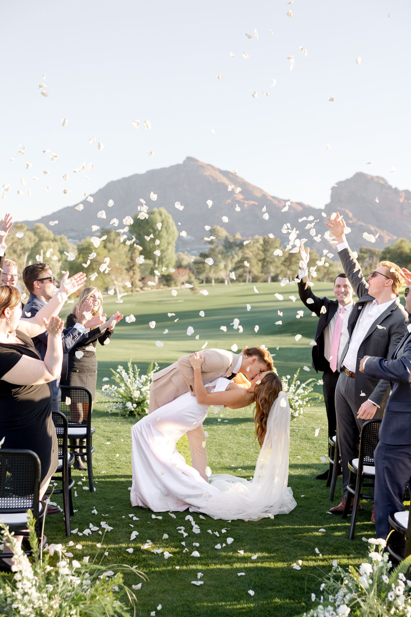 Joyful wedding ceremony outdoors with white flower petals being tossed in the air against scenic mountain backdrop.