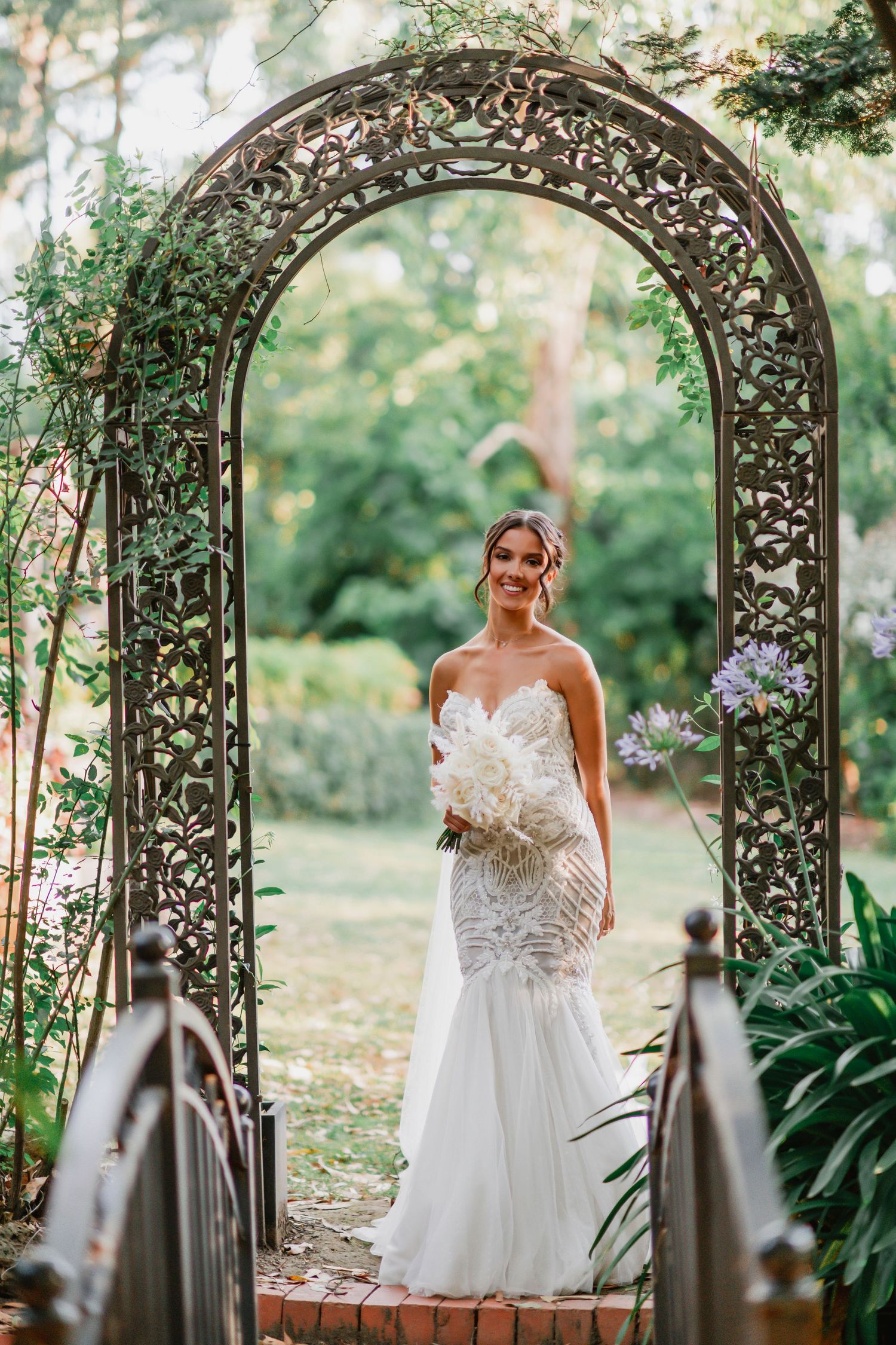 An elegant white mermaid wedding gown under an ornate garden archway decorated with greenery and vines.