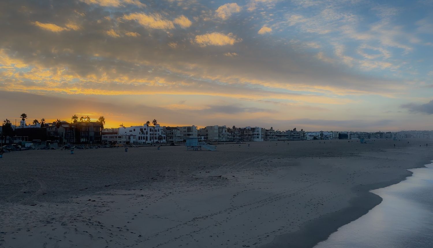 Beachfront homes and buildings line a sandy shore during a dramatic sunset with clouds casting golden light.