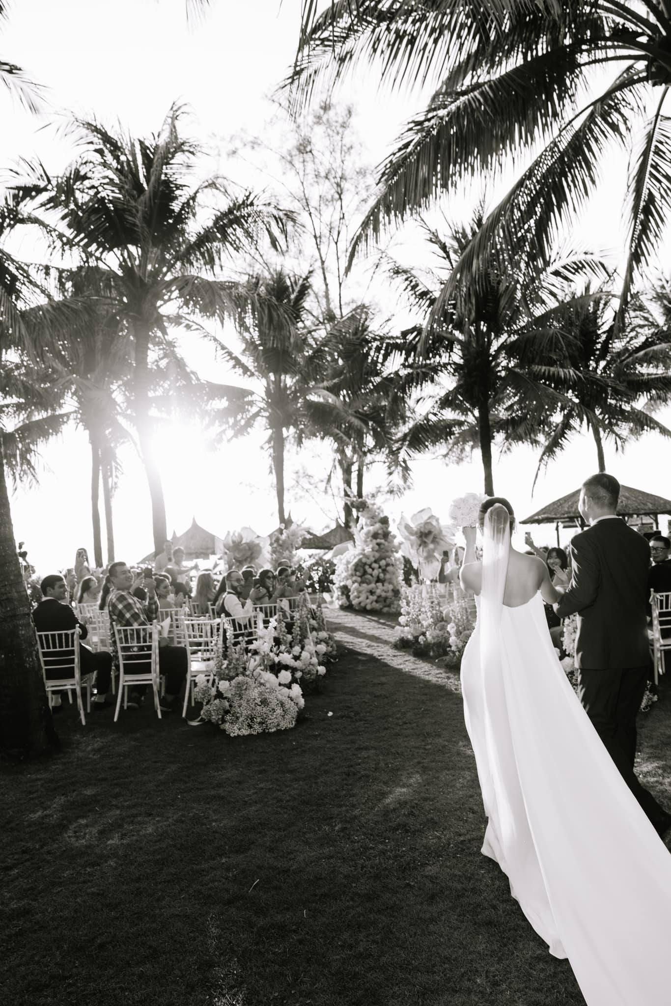 Black and white photo of bride walking toward outdoor wedding reception under tall palm trees.
