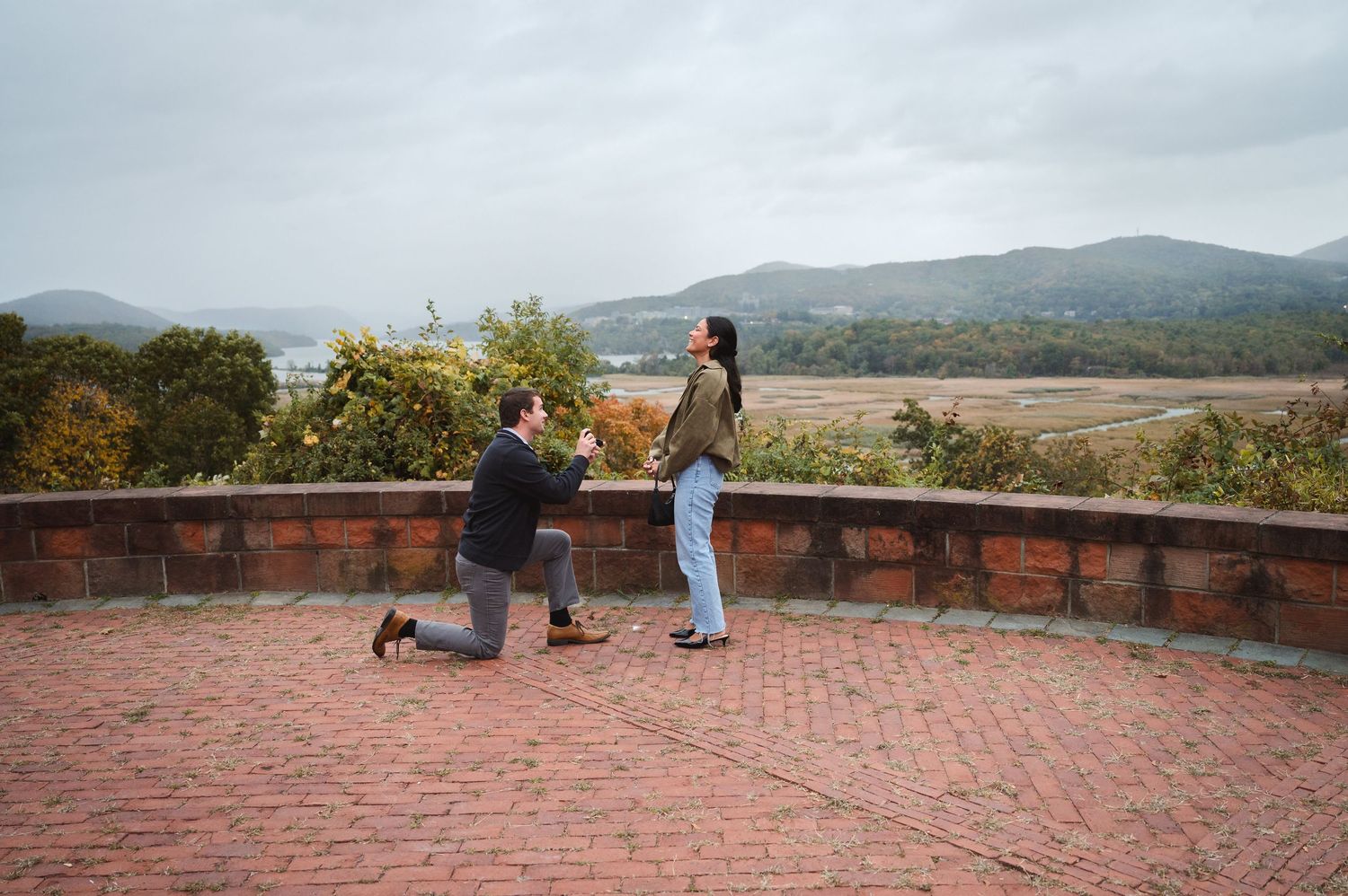 Hudson Valley wedding couple laugh and walk up hillside with fall foliage and pond behind them at Drowned Lands Brewery in Warwick, NY.