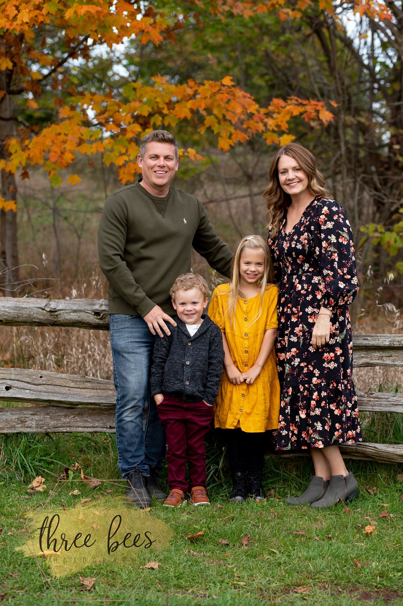 Family poses together on rustic wooden fence during autumn photoshoot with colorful orange leaves in background.