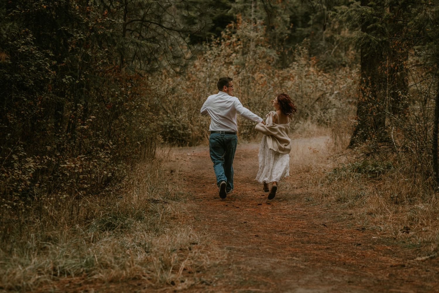 A couple walks hand in hand down a wooded path during autumn, surrounded by fallen leaves and golden light.