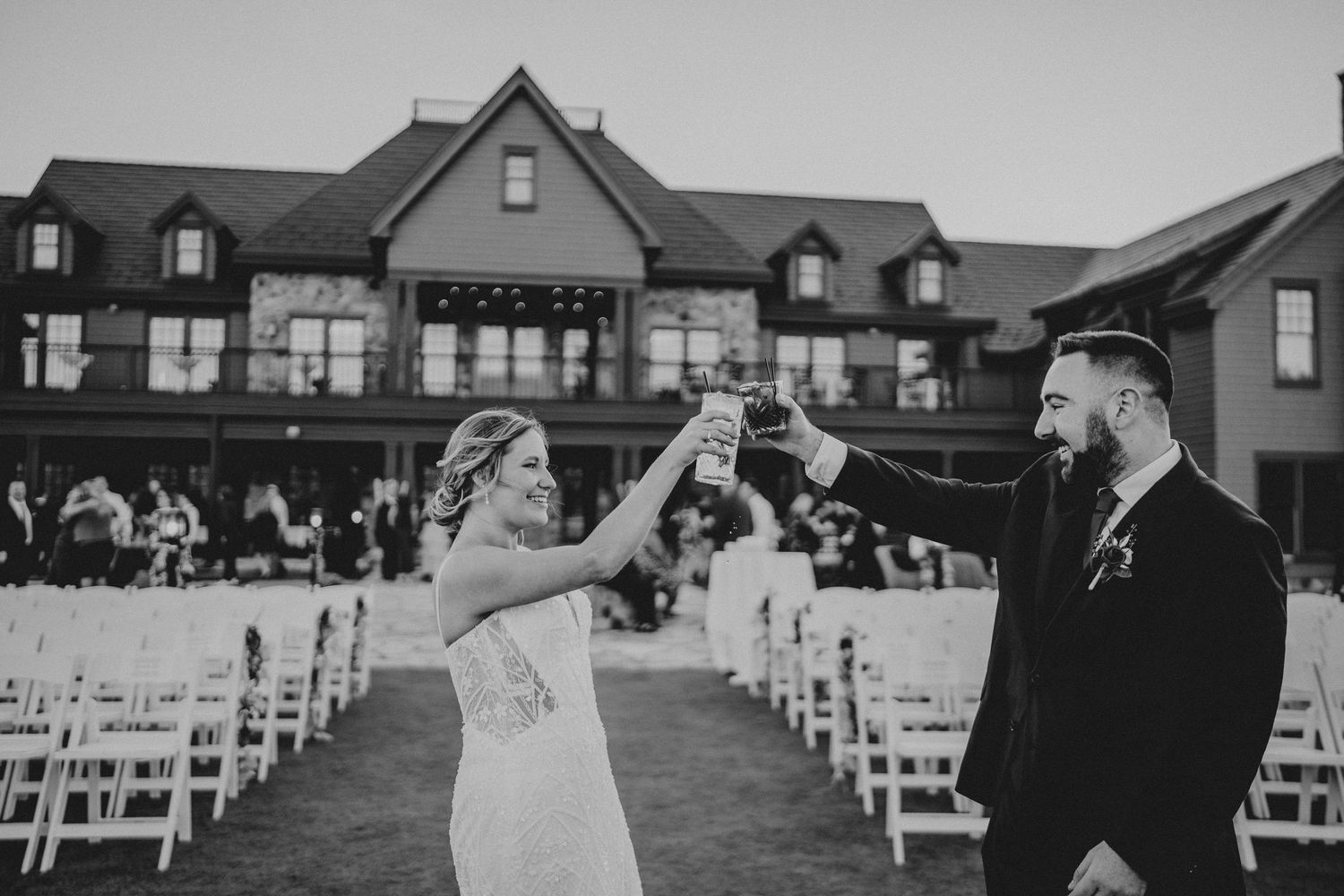A couple celebrates during an outdoor wedding ceremony with white chairs and a large mansion in the background.