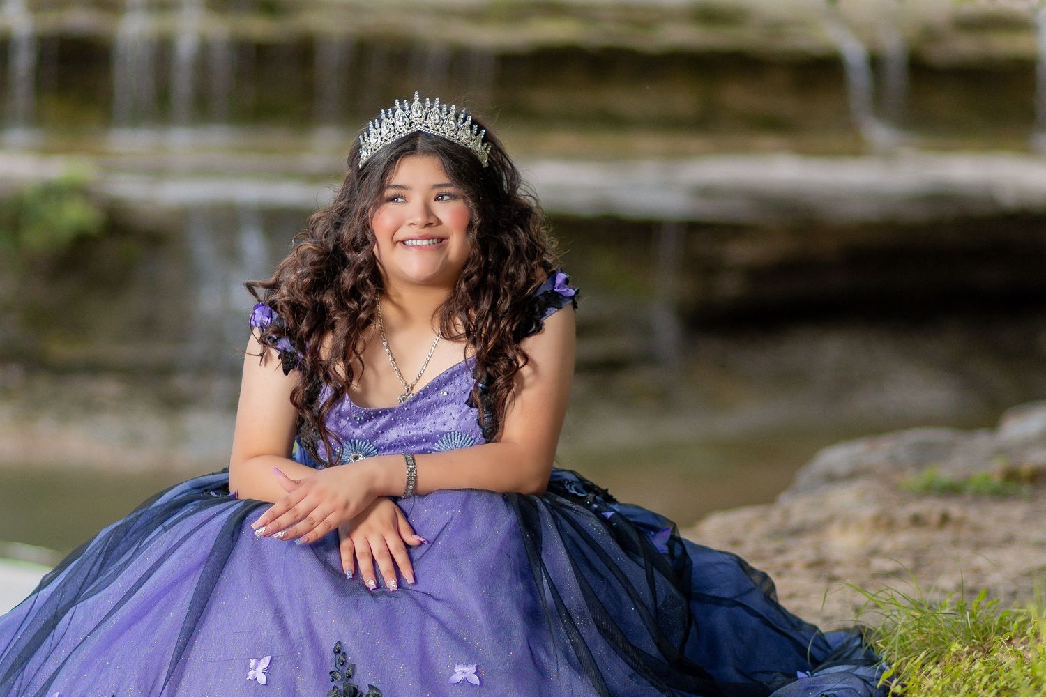 Young woman in purple quinceañera gown sitting by waterfall.