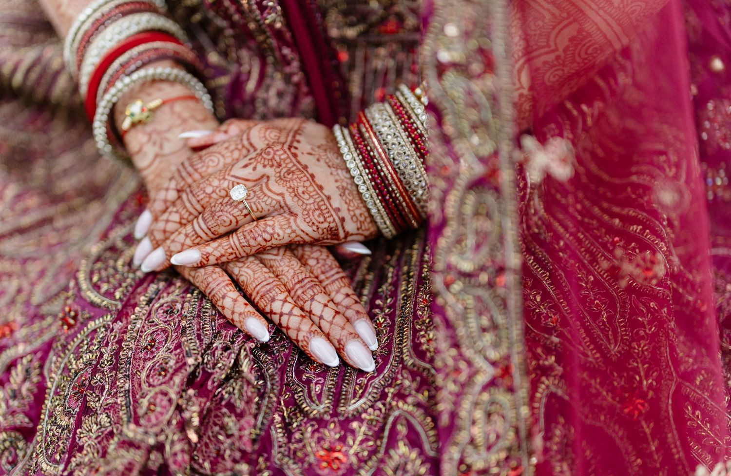 Intricate henna designs and traditional Indian bridal jewelry details shown on hands against rich red fabric.