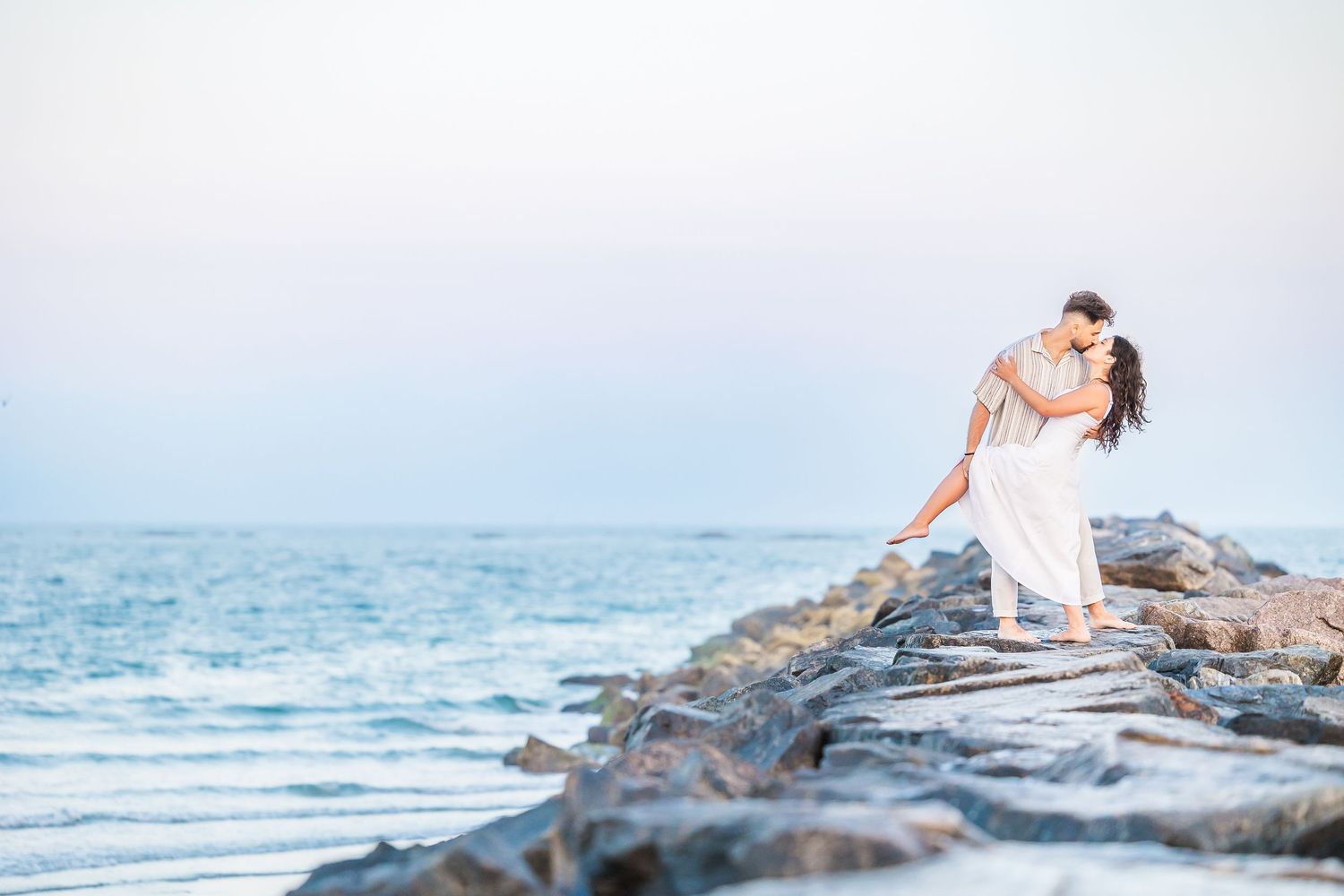 Couple in white embracing on rocky beach jetty at sunset with ocean waves in soft pastel light.