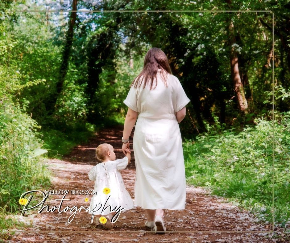 Two figures in white dresses walk hand in hand down a wooded trail surrounded by lush greenery on a summer day.