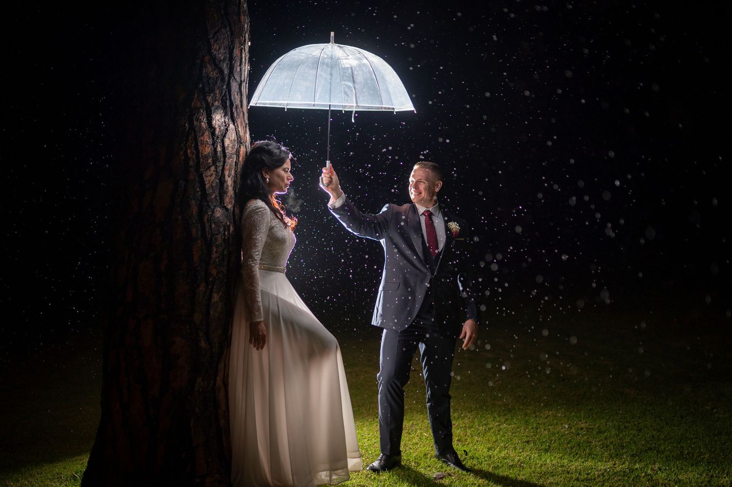 A couple in wedding attire shares a romantic moment under a clear umbrella in the rain at night.