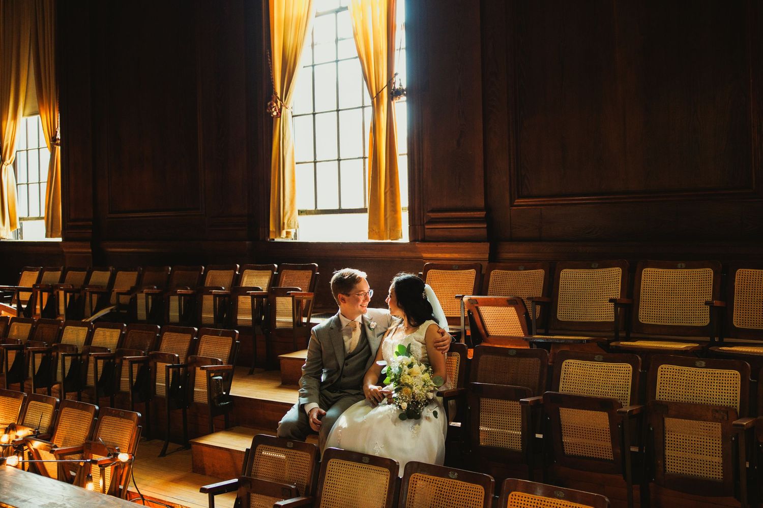 Wedding couple seated in historic wooden auditorium with sunlight streaming through tall windows.