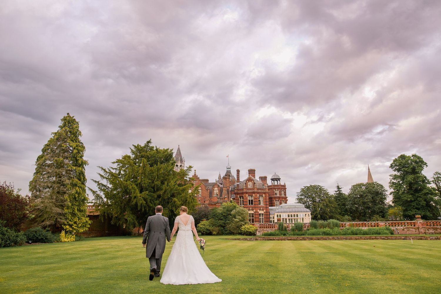 A couple walks through a scenic park with trees and a winding stream on their wedding day.