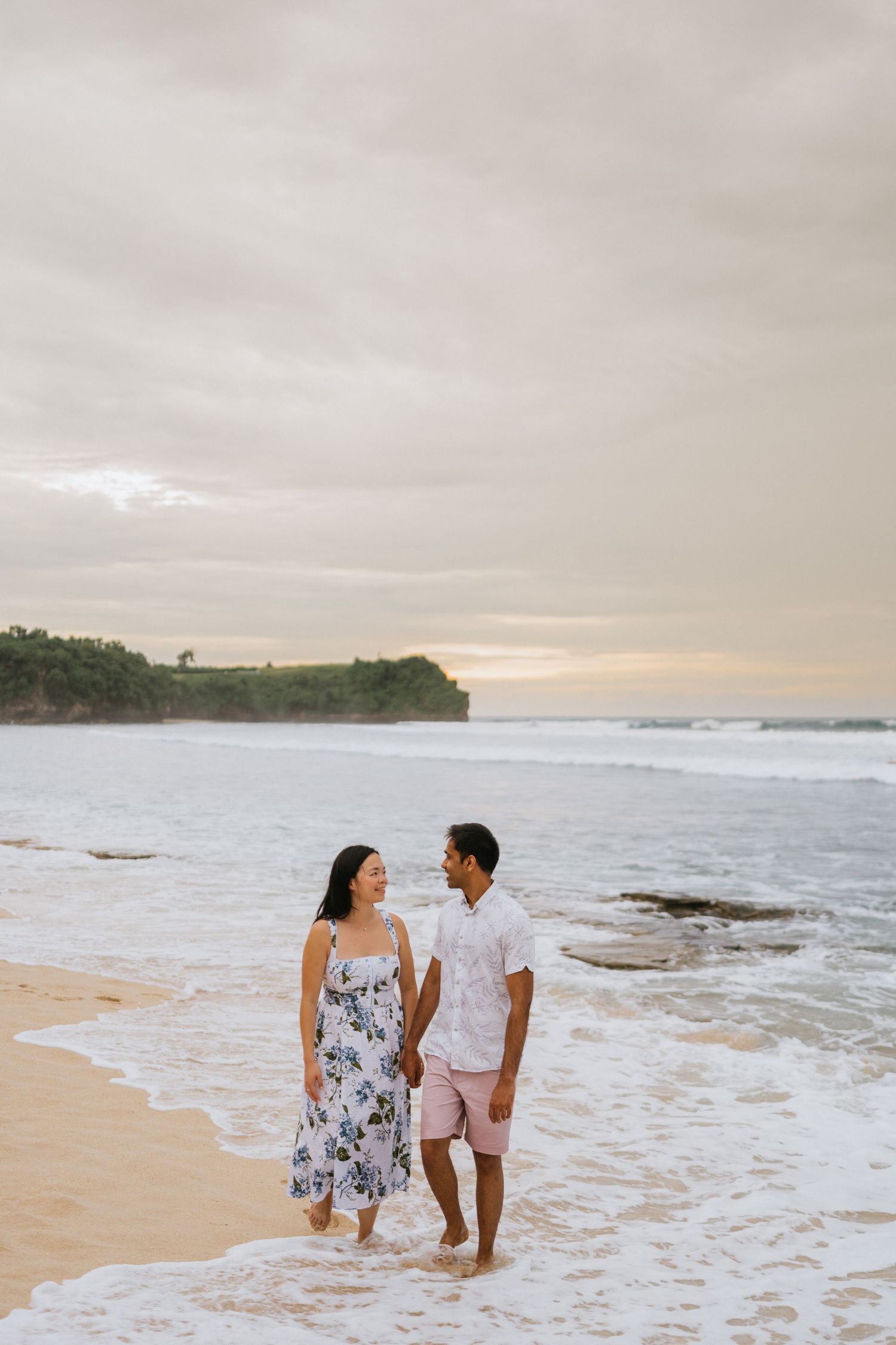 Two figures embrace on a scenic beach with dramatic rock formations and crashing waves in the background.