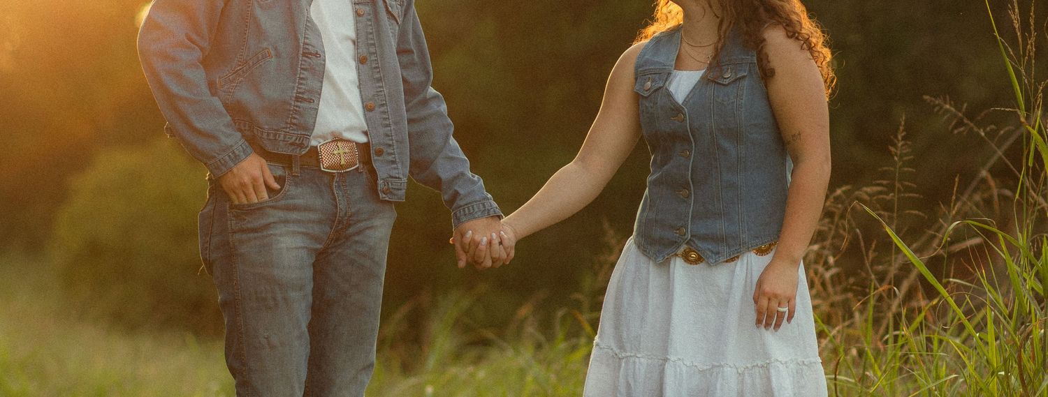 Couple in denim vests holds hands while walking through a sunlit field at golden hour.