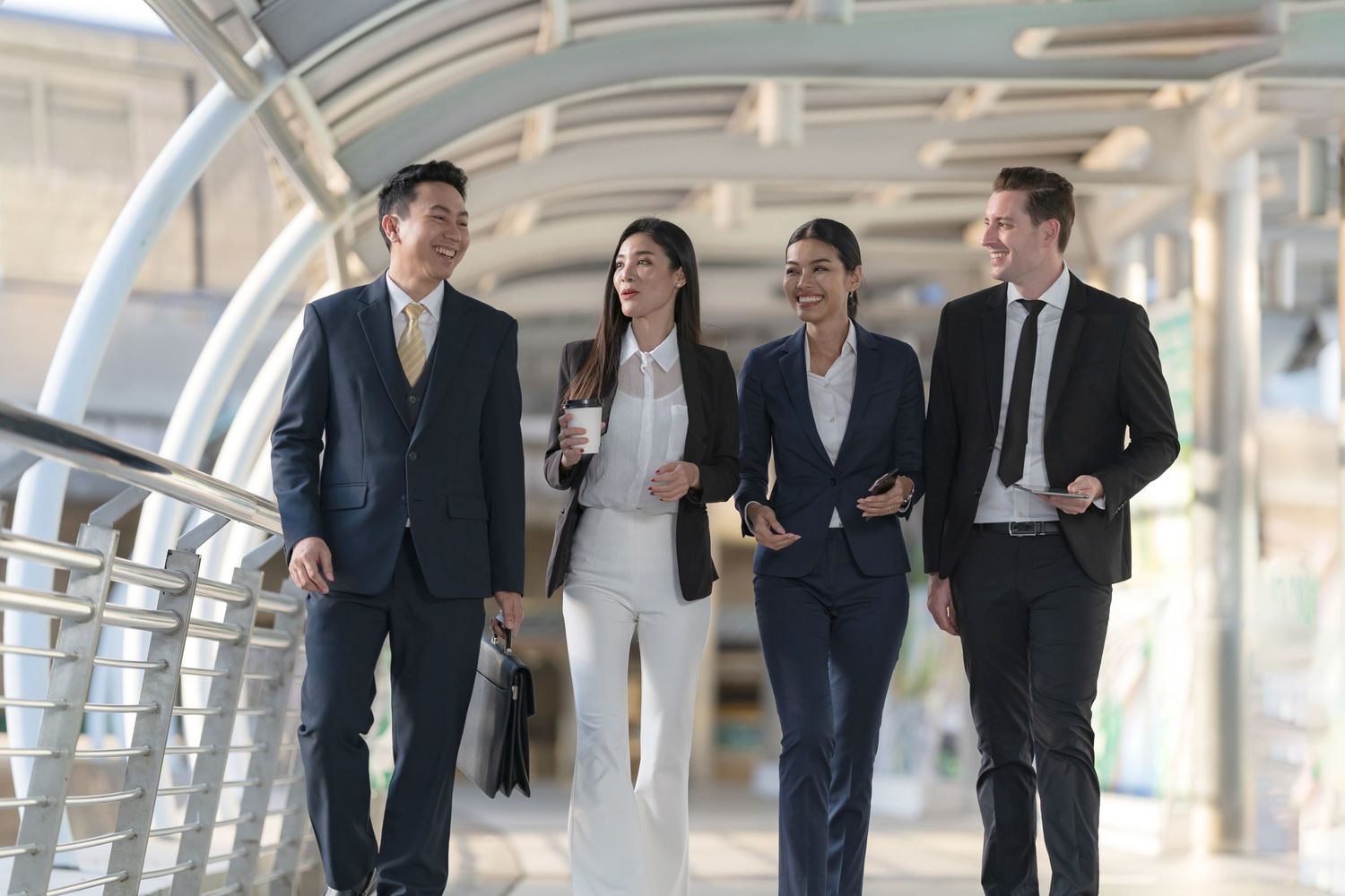 Business professionals walking and talking together in a modern office building walkway with glass walls and natural light.