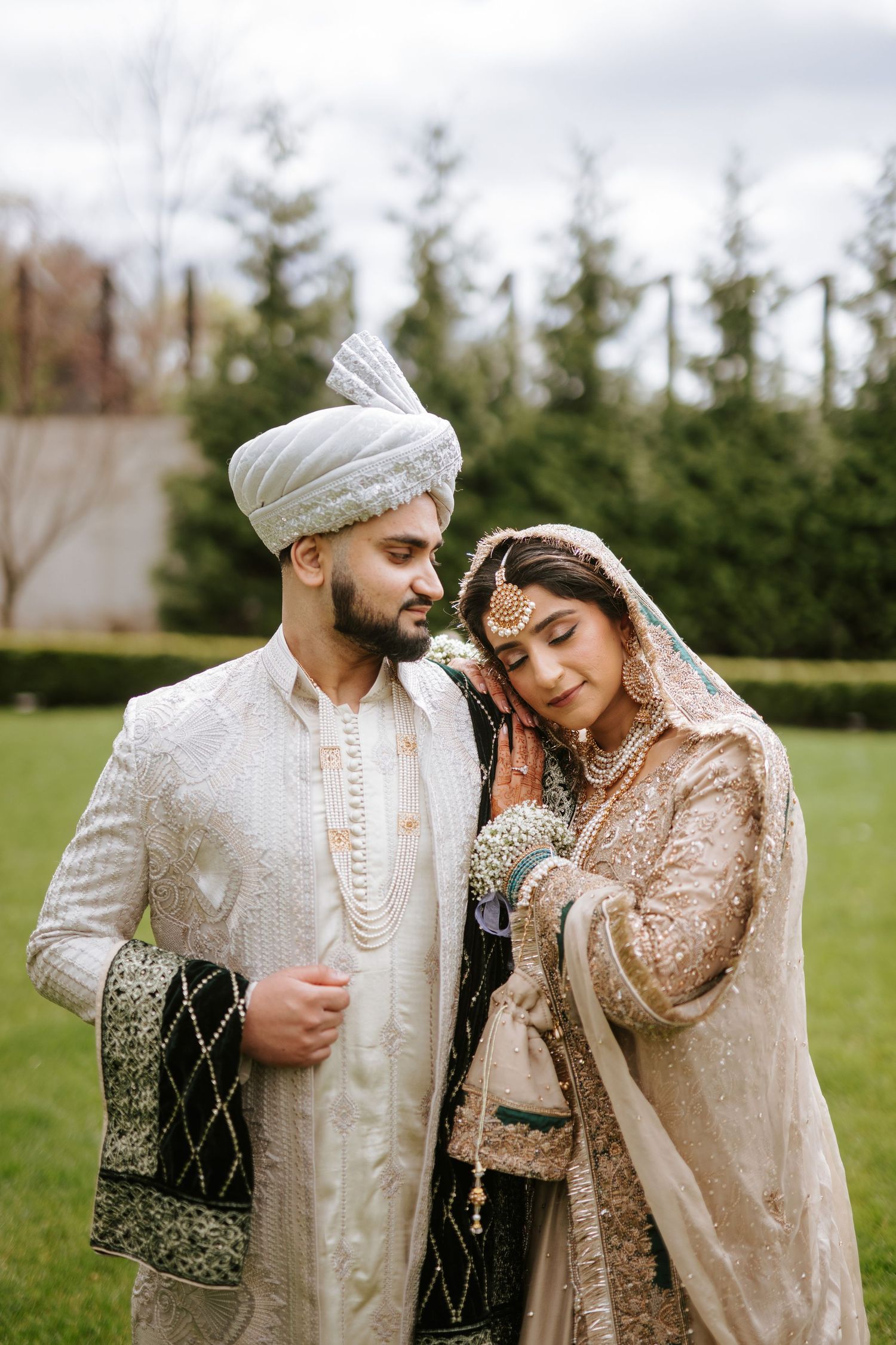 A couple in traditional South Asian wedding attire embrace outdoors on a green lawn with evergreen trees in background.