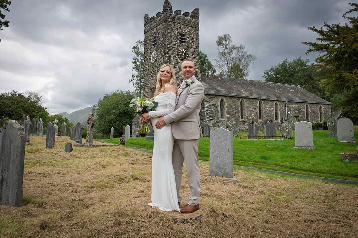 Bride and groom pose in a historic church graveyard on their wedding day, surrounded by stone tombstones.