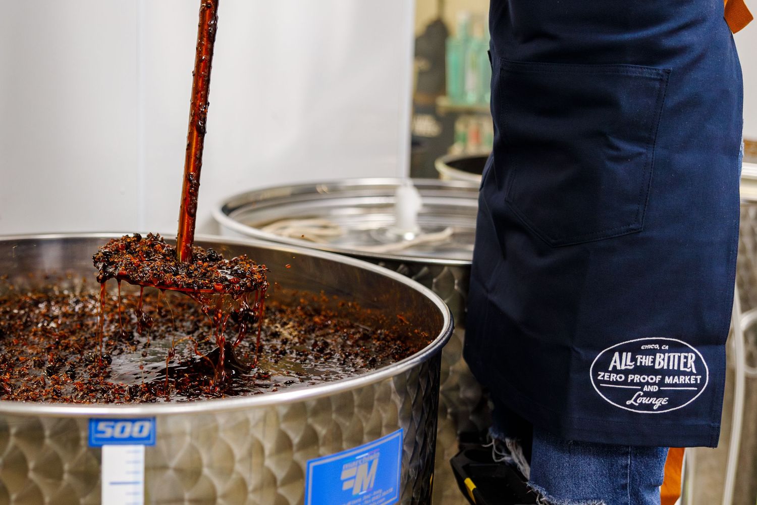 A person wearing blue overalls stirs a large metal container filled with coffee beans during the roasting process.