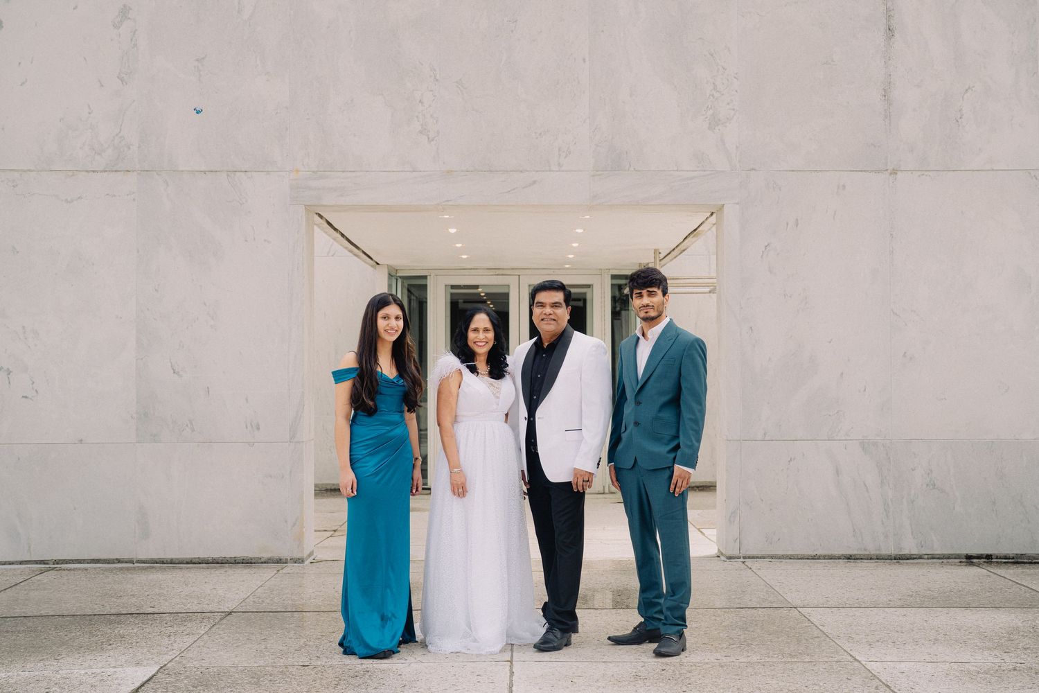 People in formal wedding attire stand against a textured concrete wall in an urban setting.