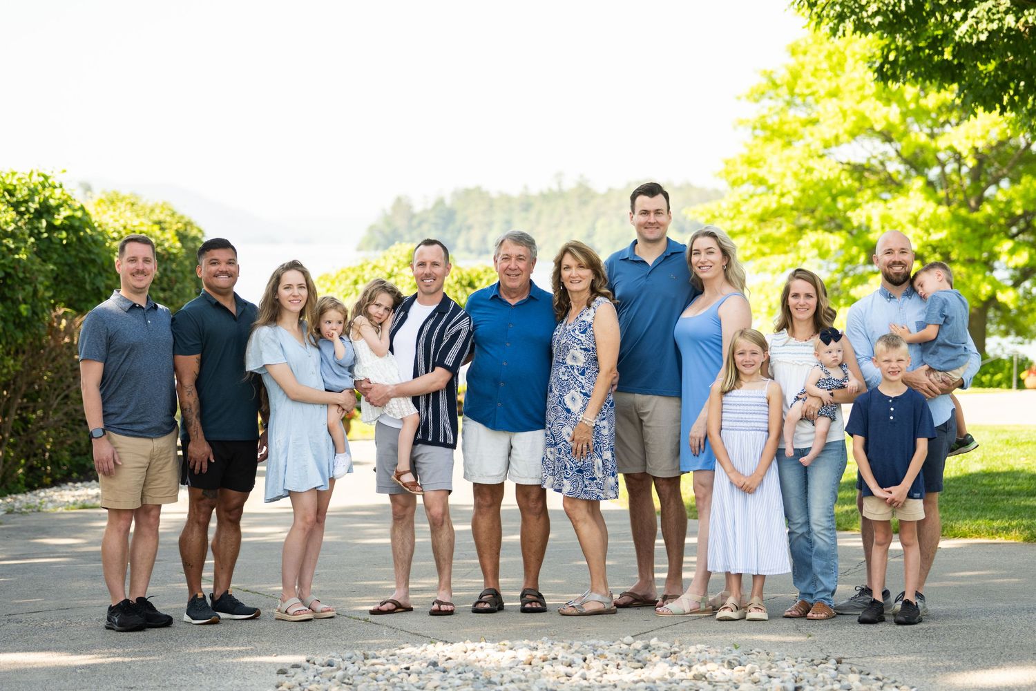 A large family group stands together outdoors on a sunny summer day with green trees and natural light in the background.
