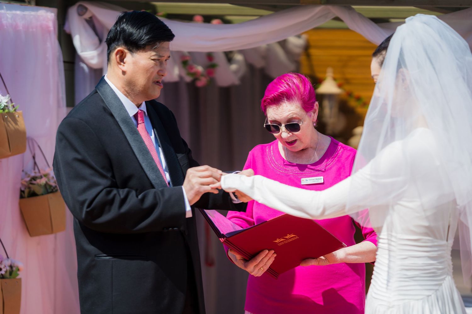A wedding officiant in a black suit and a person in bright pink attire conduct a ceremony under pink decorations.
